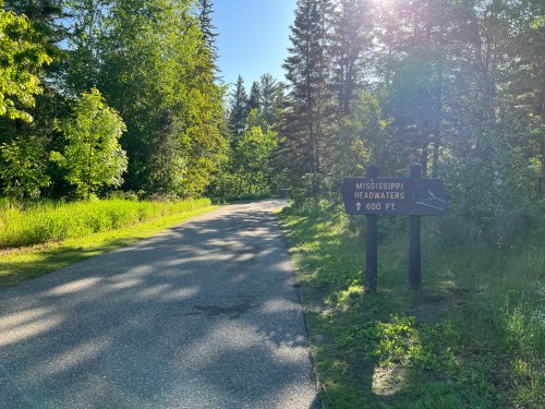 A paved path leads through lush greenery, with a sign indicating the Mississippi Headwaters is 600 feet ahead.