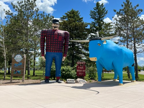 A large statue of Paul Bunyan in a plaid shirt stands next to a blue ox, with trees and a sign in the background.