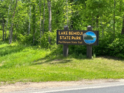Sign for Lake Bemidji State Park and Paul Bunyan State Trail, surrounded by green trees and grass.