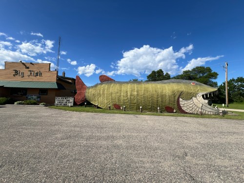 A large, whimsical fish sculpture outside a building, with blue skies and clouds in the background.