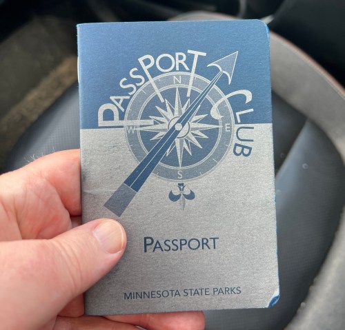 A hand holding a Minnesota State Parks Passport Club booklet featuring a compass design.