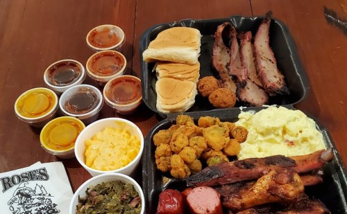 A barbecue platter featuring brisket, sausage, fried okra, macaroni and cheese, and several sauces, with dinner rolls.