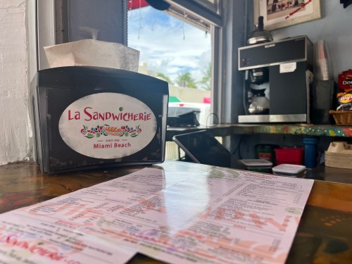 A close-up of a menu and a condiment holder at La Sandwicherie in Miami Beach, with a coffee machine in the background.