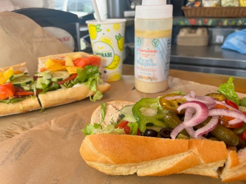 A close-up of a colorful sandwich with vegetables, alongside another sandwich and a drink in a takeout setting.