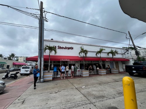 A sandwich shop with a red awning, outdoor seating, and people gathered outside on a cloudy day.