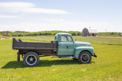 A vintage teal truck parked on green grass with a barn in the background under a clear blue sky.