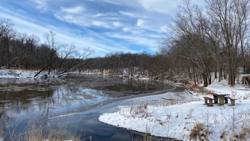 A serene winter scene of a river with ice, surrounded by snow-covered banks and bare trees under a cloudy sky.