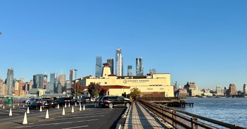A waterfront view of a city skyline with modern skyscrapers, a restaurant, and parked cars along a pier.