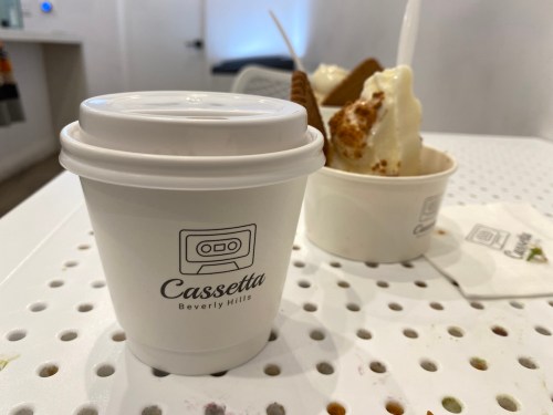 A white coffee cup with a logo sits on a table, with a dessert in the background.