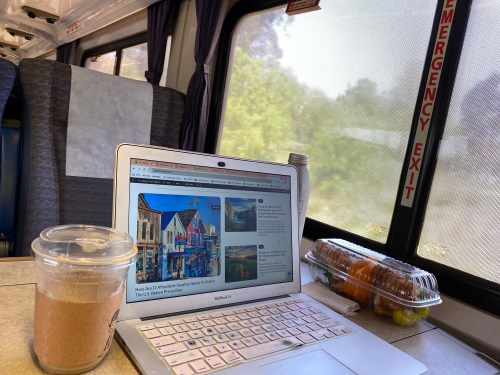 A laptop on a train table with a drink and snacks, showing a travel website on the screen. Sunlight through the window.