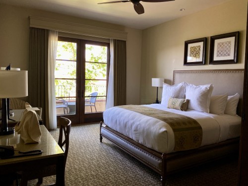 A cozy hotel room featuring a king-sized bed, desk, and balcony access with natural light streaming in.