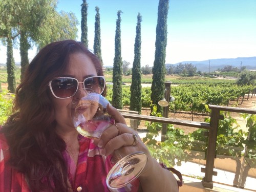 A woman in sunglasses enjoys a glass of wine, surrounded by vineyards and tall trees under a clear blue sky.