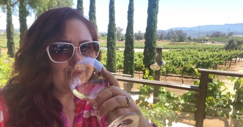 A woman in sunglasses enjoys a glass of wine, with vineyards and tall trees in the background under a clear blue sky.