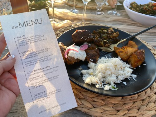 A hand holds a menu beside a plate of food featuring rice, meat, potatoes, and greens, set on a table outdoors.