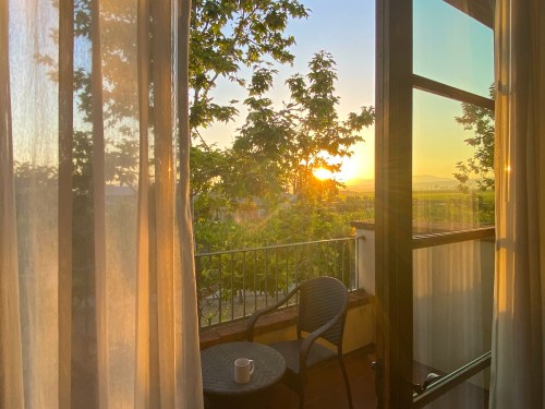 A sunlit balcony with a chair and a cup, framed by sheer curtains and greenery, overlooking a scenic landscape.