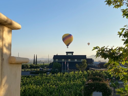 A colorful hot air balloon floats over a vineyard and a building under a clear blue sky.