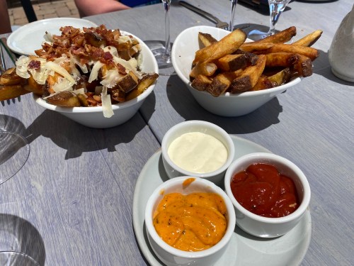 Two bowls of fries, one topped with bacon and cheese, accompanied by three dipping sauces: ketchup, creamy sauce, and orange sauce.