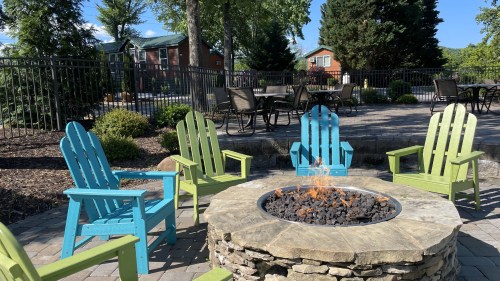 A cozy outdoor fire pit surrounded by colorful Adirondack chairs in a landscaped area with trees and buildings in the background.