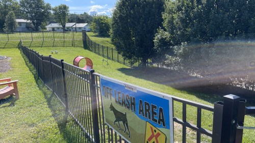 A fenced off-leash dog area with a sign, grassy space, and agility equipment in the background.