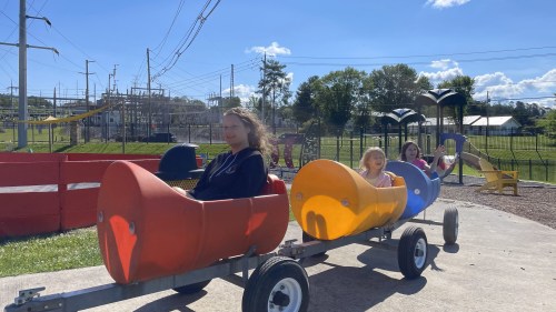 Three children ride a colorful train on a sunny day at a park, with power lines and trees in the background.