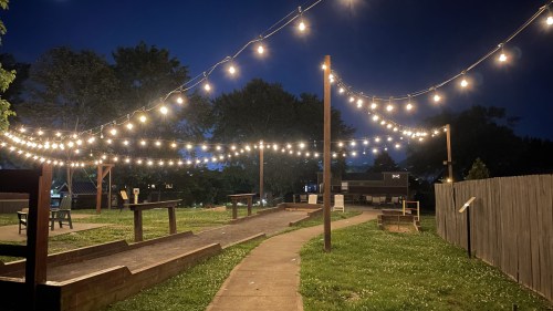 A pathway illuminated by string lights in a park at night, with seating areas and greenery in the background.