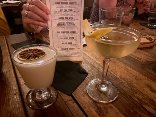 Two cocktails on a wooden table: one with a dried lemon slice and the other garnished with a lemon twist, alongside a menu.