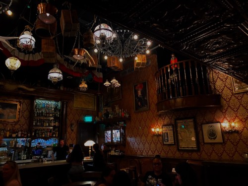 A cozy bar interior with vintage decor, chandeliers, and a wooden balcony, featuring patrons enjoying their drinks.