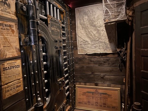 A dimly lit room featuring a large vault door, old maps, and vintage posters on wooden walls.