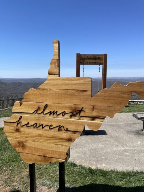 Wooden sign shaped like West Virginia with the text "almost heaven" against a clear blue sky and distant mountains.