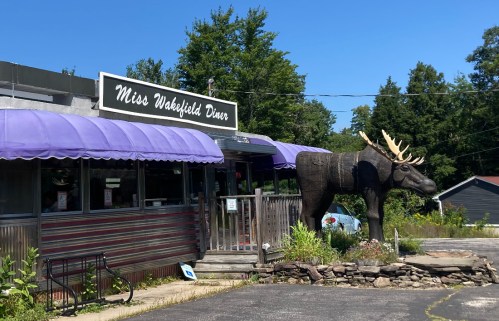 A diner with a purple awning and a large moose statue outside, surrounded by trees and a clear blue sky.