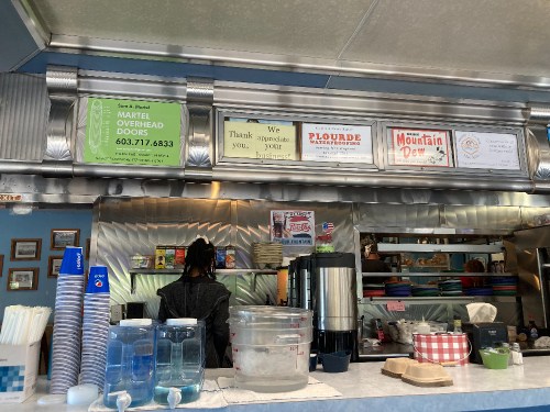 A diner counter with signs, a coffee pot, and a person behind the counter preparing food.
