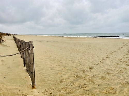 A sandy beach with a wooden fence, overcast skies, and gentle waves in the distance.