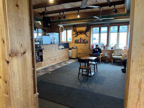 Cozy interior of a rustic café with wooden decor, tables, and a view of the outdoors. Two people are seated.