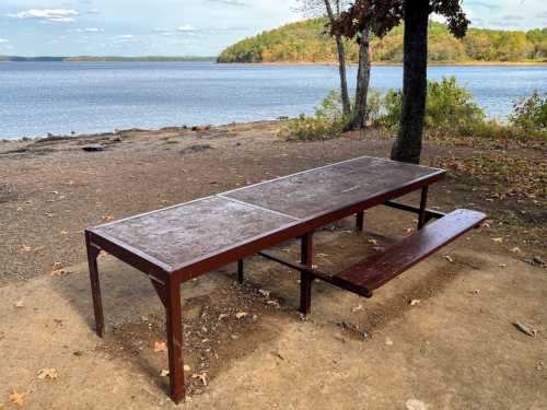 A picnic table by a calm lake, surrounded by trees and autumn foliage under a clear blue sky.