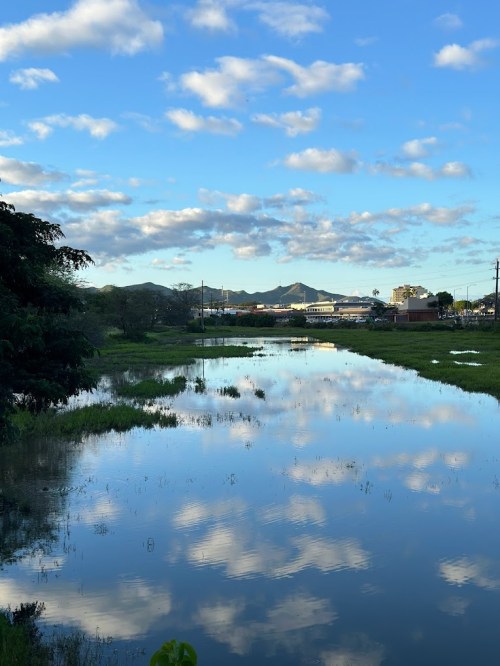 A serene landscape featuring a calm river reflecting clouds and mountains under a blue sky. Lush greenery surrounds the water.