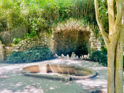 A stone archway surrounded by greenery, with a shallow stone basin in the foreground and rocky formations nearby.