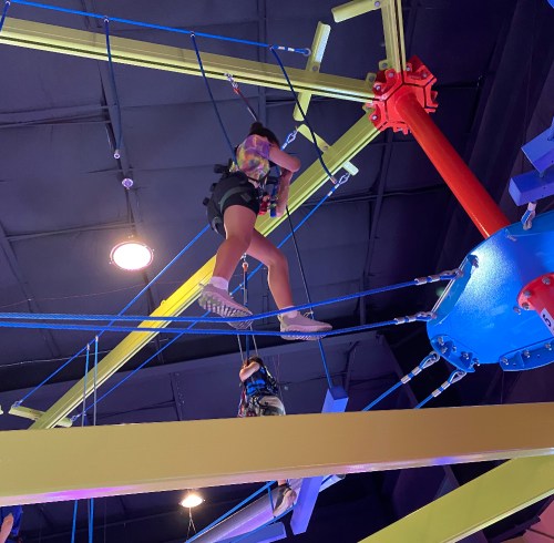 A person climbs on a colorful indoor ropes course, suspended high above the ground.