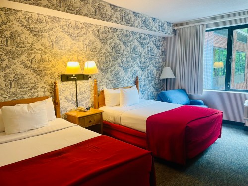 A cozy hotel room with two beds, a nightstand, a lamp, and a window with natural light. Decorated with patterned wallpaper.