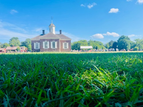A historic building with a dome, surrounded by green grass and trees, under a clear blue sky with fluffy clouds.