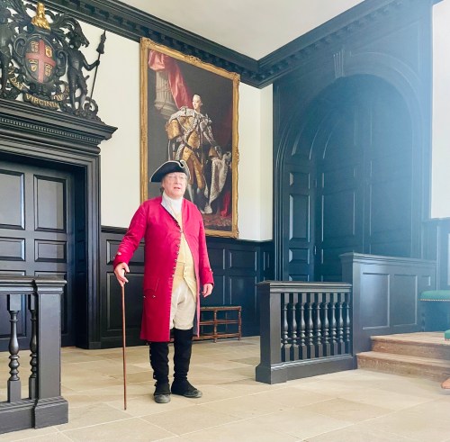 A man in historical attire stands in a dark-paneled room with a large portrait and wooden staircase.
