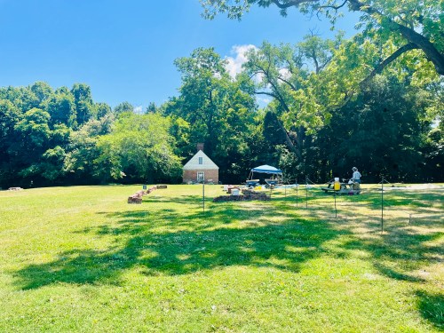 A sunny outdoor scene featuring a grassy area with a small house, trees, and a tent set up for an event or activity.