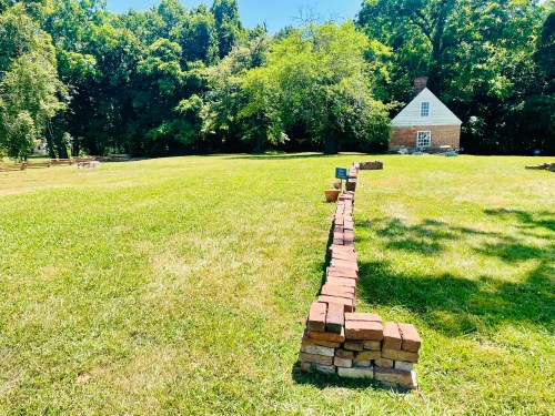 A grassy field with a brick wall leading to a small, white house surrounded by trees.