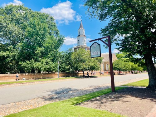 A sunny street lined with trees, featuring a church steeple and a sign, with people walking and a dog in the distance.
