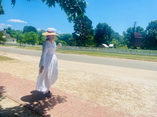A person in a white dress and straw hat walks along a sunny path with trees and a windmill in the background.