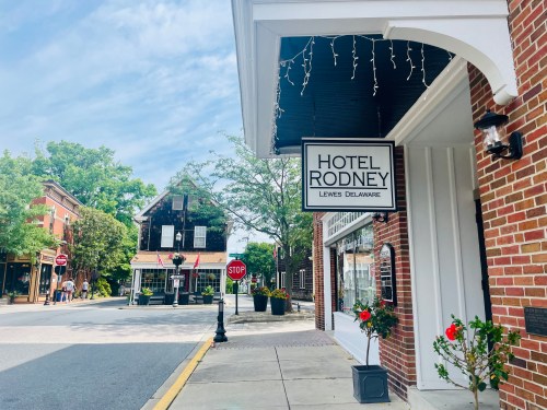 Sign for Hotel Rodney in Lewes, Delaware, with a charming street view and greenery in the background.