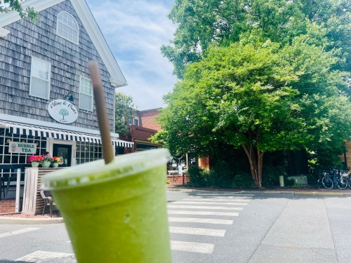 A green smoothie in a cup held in front of a tree-lined street with shops and a bubble tea store in the background.