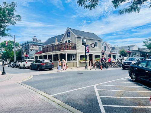 A charming street scene featuring a restaurant with outdoor seating, cars parked, and people walking in a quaint town.