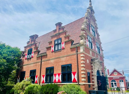 Historic brick building with a steep roof, decorative windows, and red and white banners, surrounded by greenery.