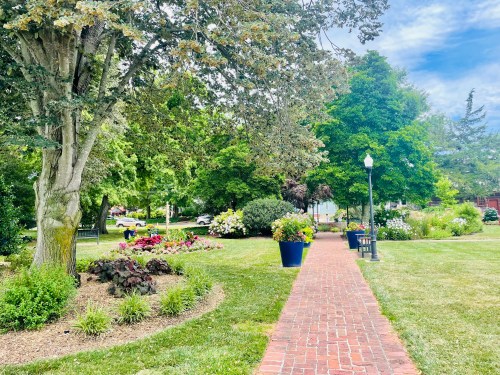A brick path winds through a lush park with colorful flowers, trees, and benches under a bright blue sky.