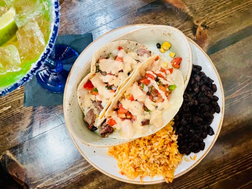 Two tacos filled with meat and toppings, served with black beans and rice on a wooden table, alongside a drink.